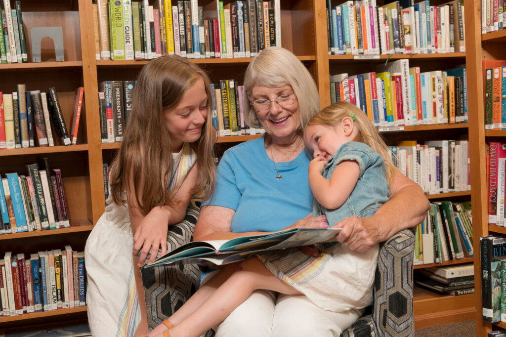 A senior woman sits in a chair in a library and reads a book to her two granddaughters