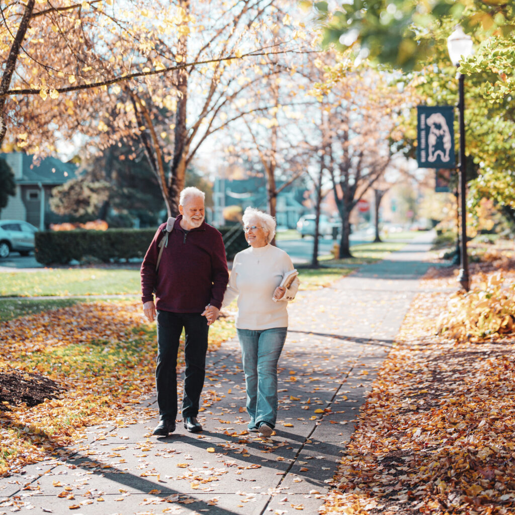A senior couple walks down a sidewalk among autumn leaves